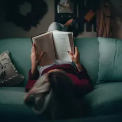 Osoba leżąca na kanapie czyta otwartą książkę.