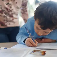 Chłopiec pisze w zeszycie, a obok niego stoi osoba w bluzce w kwiaty.