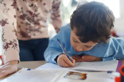 Chłopiec pisze w zeszycie, a obok niego stoi osoba w bluzce w kwiaty.